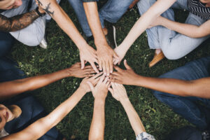 Grupo de pessoas coloca as mãos uma sobre as outras em um parque. Passa uma mensagem de união.